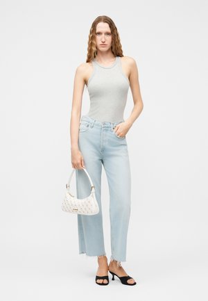 Γυναίκα με μακριά σγουρά μαλλιά, ντυμένη με αμάνικο τοπ ανοιχτού γκρι χρώματος, ανοιχτόχρωμο μπλε τζιν, μαύρα σανδάλια με τακούνι και κρατάει μια λευκή τσάντα χειρός.