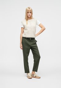 Beige színű kötött ujjatlan felső fehér póló fölött, olívazöld nadrággal és mintás barnás tornacipővel párosítva. Egyszerű kialakítás, semleges árnyalatokkal.