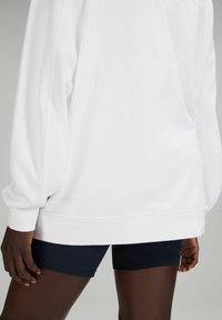 Felpa bianca oversize con maniche lunghe, polsini e vita a costine. Indossata sopra a pantaloni corti blu navy. Texture liscia e vestibilità comoda.