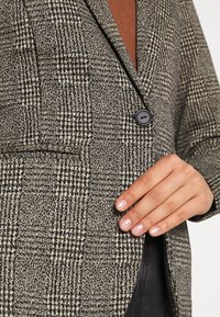 Main reposant sur un blazer à carreaux noir et blanc avec un bouton, par-dessus un haut marron.