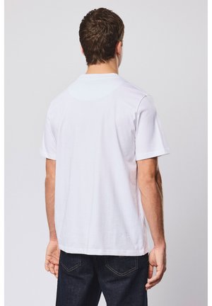 Mann mit kurzem braunem Haar, der ein einfaches weißes T-Shirt und dunkle Jeans trägt, steht mit dem Rücken zum Betrachter vor einem schlichten hellen Hintergrund.