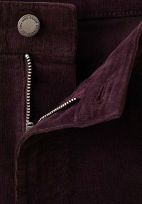 Pantaloni din catifea mov închis, prim-plan care arată fermoarul metalic argintiu parțial deschis și nasturele argintiu gravat cu textul brandului.