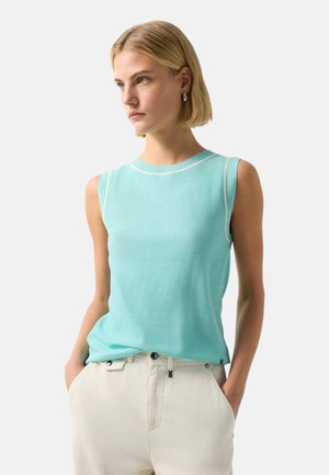 Vrouw met een blonde bobkapsel, draagt een mouwloos lichtblauw topje en gebroken witte broek, handen in de zakken, kijkt opzij tegen een effen achtergrond.