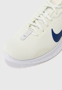 Zapatilla deportiva de color claro con una parte superior texturizada, cordones blancos y un prominente logo de swoosh azul de Nike en el lateral.