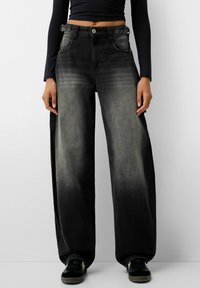 Črne široke jeans hlače iz denima z zbledelim gradientnim učinkom, opremljene s sprednjimi žepi in gumbom. Nosijo se s črnimi supergami.