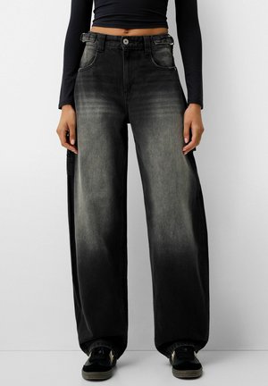Jeans a gamba larga in denim nero con effetto sfumato, dotati di tasche frontali e chiusura con bottone. Indossati con scarpe sportive nere.