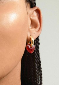 Gouden oorring met een rood hartvormig hangertje, met een gladde textuur en glanzende afwerking, gedragen in een vlechten kapsel.