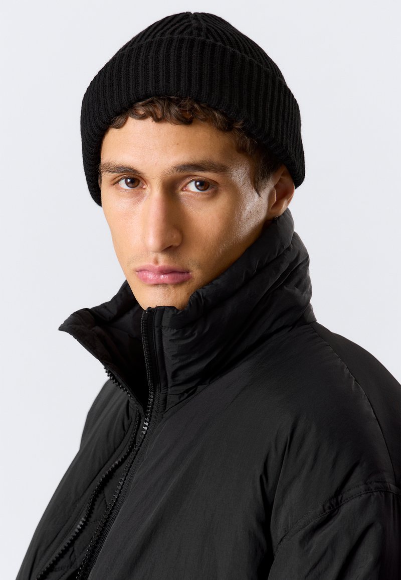 Chaqueta acolchada negra con cuello alto y cierre de cremallera, combinada con un gorro de canalé negro. El modelo muestra una expresión neutral frente a un fondo liso.