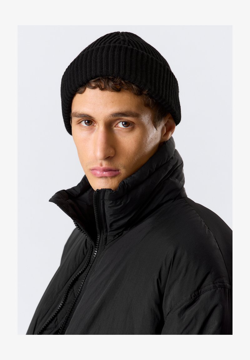 Chaqueta acolchada negra con cuello alto y cierre de cremallera, combinada con un gorro de canalé negro. El modelo muestra una expresión neutral frente a un fondo liso.