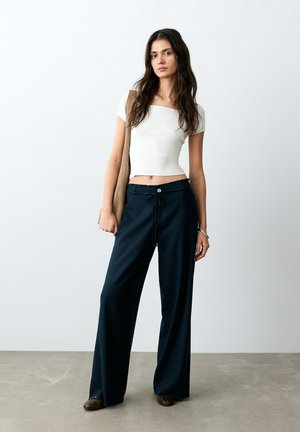 Giovane donna con lunghi capelli scuri, indossa un top bianco corto, pantaloni blu navy a gamba larga, scarpe marroni e porta una borsa a mano beige.