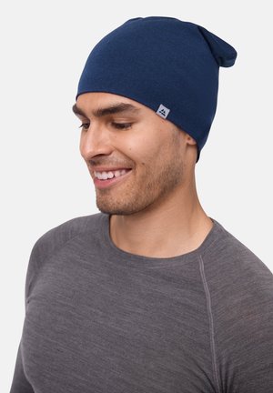 Bonnet en tricot bleu marine avec un sommet effilé et une texture subtile. Présente une petite étiquette rectangulaire sur le côté. Porté avec une chemise à manches longues grise.