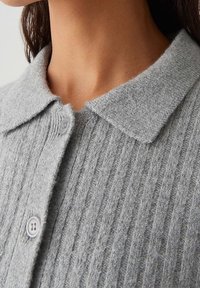 Cardigan gris en tricot avec une texture côtelée, col plat et fermeture à deux boutons. Le tissu semble doux et confortable, mettant en valeur les détails du tricot.