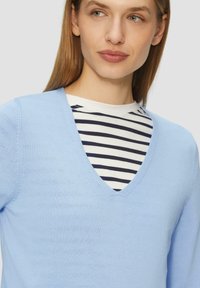 Helles blaues V-Ausschnitt-Pullover aus weichem Stoff, drüber getragen über einem weiß-blau gestreiften Hemd. Hat lange Ärmel und eine figurbetonte Passform.