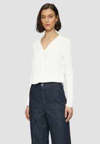White long-sleeve top with a V-neck and a subtle front placket, paired with dark denim trousers featuring a side logo detail.