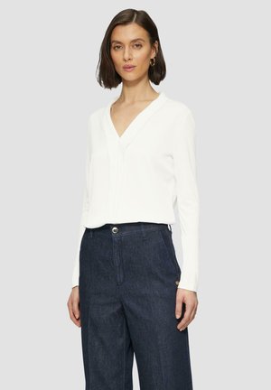 White long-sleeve top with a V-neck and a subtle front placket, paired with dark denim trousers featuring a side logo detail.