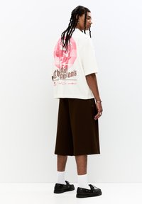 Cream oversized t-shirt featuring a pink graphic print on the back, paired with brown shorts and black shoes. White socks complete the outfit.