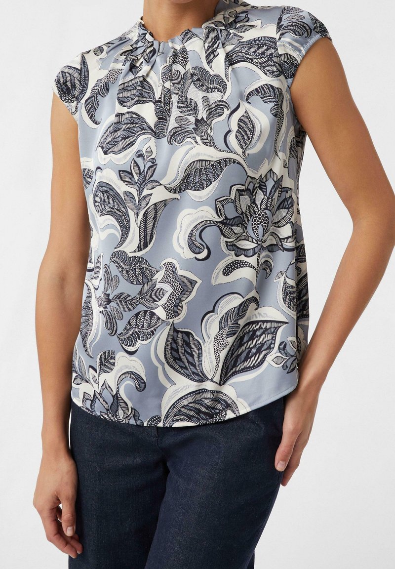 Blouse sans manches bleu pâle avec de grands motifs floraux noirs et blancs détaillés, portée avec un jean foncé, mains détendues le long du corps.