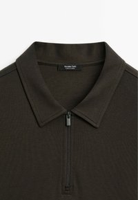 Polo di colore marrone scuro con colletto con zip, maniche corte, texture liscia e un'etichetta con la scritta "Massimo Dutti". Prodotto in Turchia.