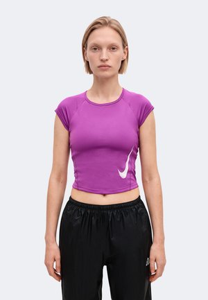 Une femme porte un crop top Nike violet orné d’un Swoosh blanc et un pantalon de sport noir sur un fond clair uni.