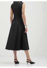 Abito in satin nero con corpetto aderente e orlo svasato. Design senza maniche, vita visibile e texture liscia. Indossato con scarpe con tacco nere.