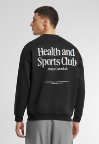 Černá mikina vyrobená z bavlny, na zádech s výrazným bílým textem: "Zdravotní a sportovní klub" a menším textem níže. Ležérní design.