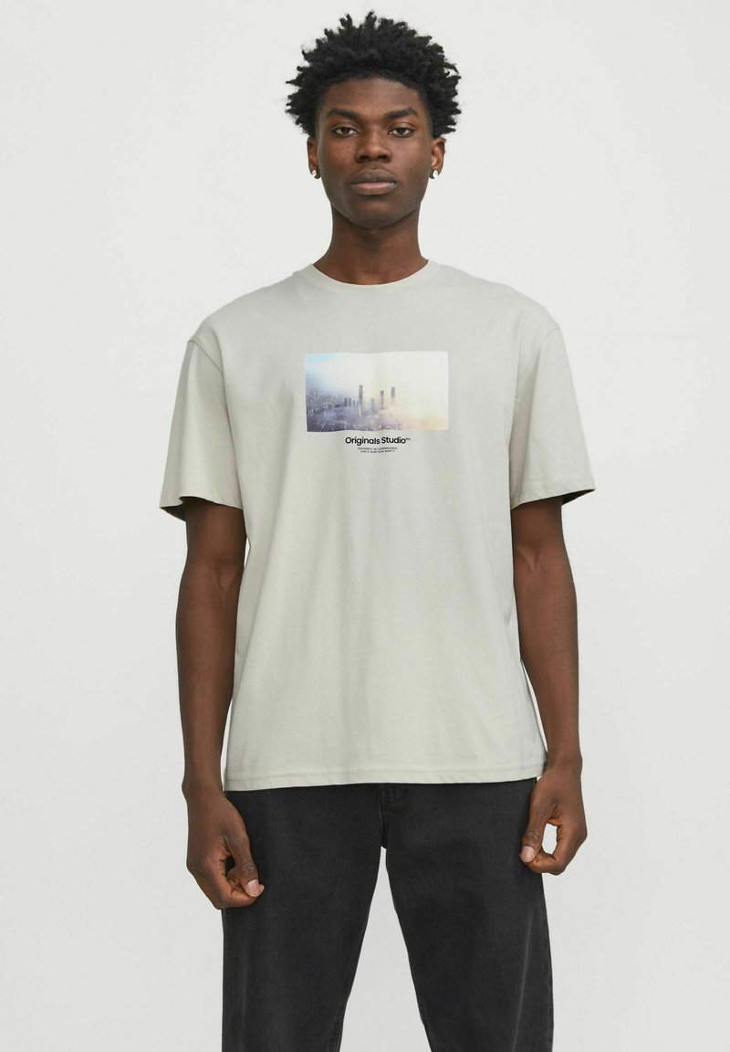 Camiseta de algodón gris claro con un estampado gráfico rectangular que presenta un paisaje urbano y el texto "Originals Studio" en el centro. Mangas cortas.