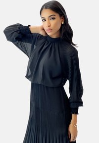 Vestido negro de plisado con mangas largas de organza y abullonadas. El escote alto y la tela texturizada crean una silueta sofisticada. Los pendientes dorados añaden un toque de detalle.