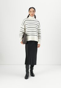 Übergroßer cremfarbener Pullover mit schwarzen Streifen und hohem Kragen, kombiniert mit einem schwarzen Satin-Midirock und kniehohen schwarzen Stiefeln. Hält eine graue Clutch.