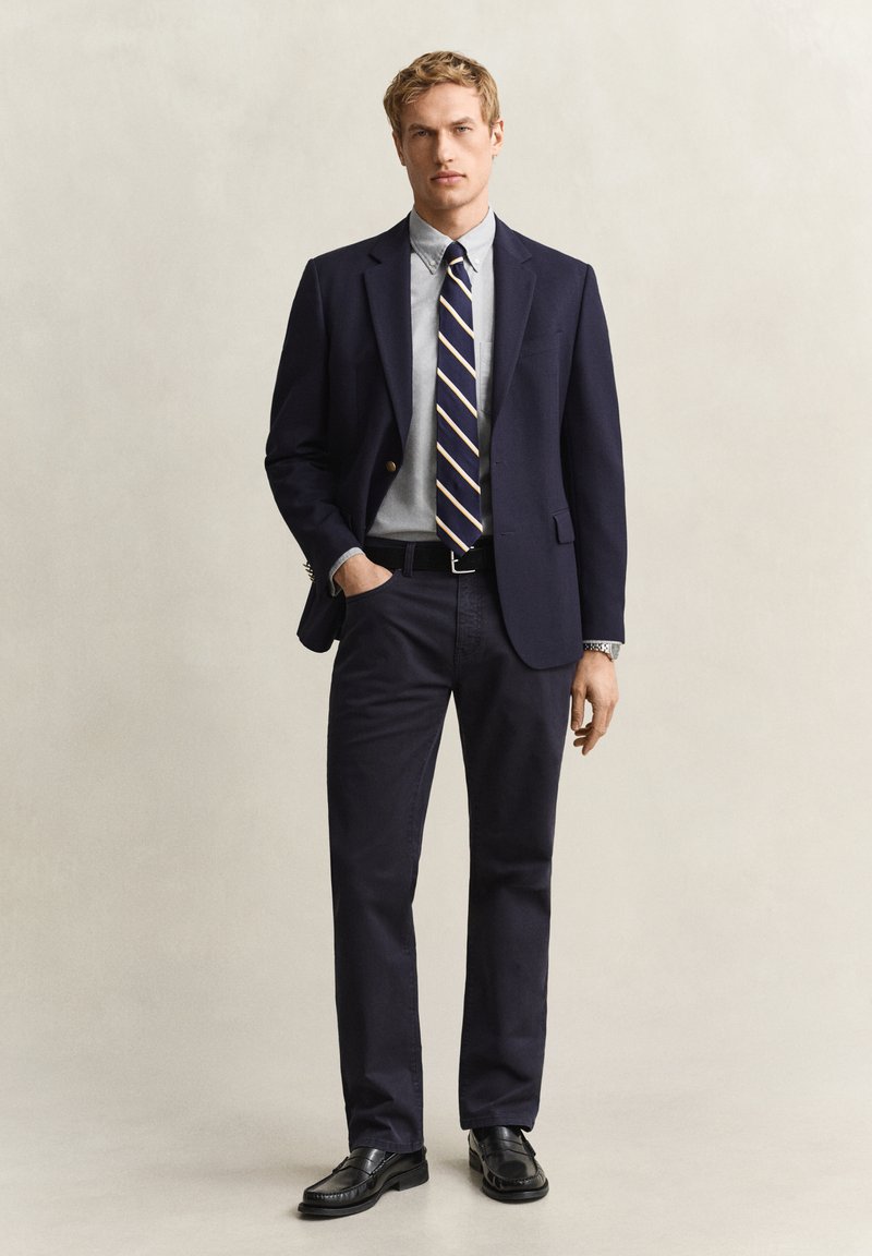 Homme blond portant un blazer bleu marine, une chemise bleu clair, une cravate rayée bleu marine, un pantalon sombre, des chaussures noires, debout avec une main dans la poche sur un fond uni.