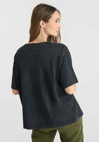 Mujer con cabello largo castaño que lleva una camisa negra de manga corta holgada y pantalones verdes, de espaldas, mirando de lado contra un fondo liso.