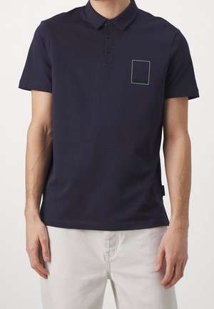 Polo de algodón azul marino con cuello con solapa, tapeta de tres botones y contorno de bolsillo cuadrado en el pecho. Mangas cortas, ajuste holgado.