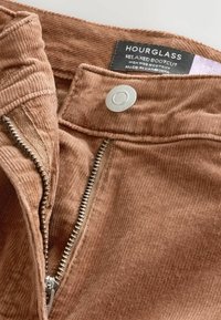 Pantalones bootcut relajados en pana marrón. Presentan un cierre frontal con cremallera y botón a presión, con una textura suave y un patrón de canalé visible.
