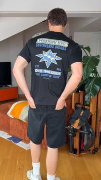 Άτομο στέκεται εσωτερικά με τα χέρια στις πίσω τσέπες, φοράει μαύρο μπλουζάκι με σχέδιο και σορτς, κοντά σε κωπηλατικό μηχάνημα και ένα μεγάλο φυτό.