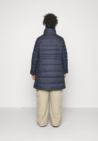 Marineblauer Steppmantel mit hohem Kragen, horizontaler Steppung, langen Ärmeln und knielangem Schnitt. Kombiniert mit hellfarbigen, locker sitzenden Hosen.