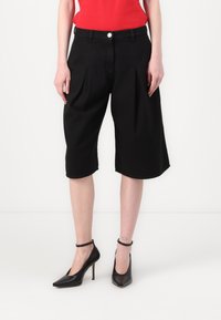 Zwarte capri-broek met geplooide voorkant, zijzakken en knoopsluiting, gecombineerd met zwarte hoge hakken. Glad stofoppervlak.