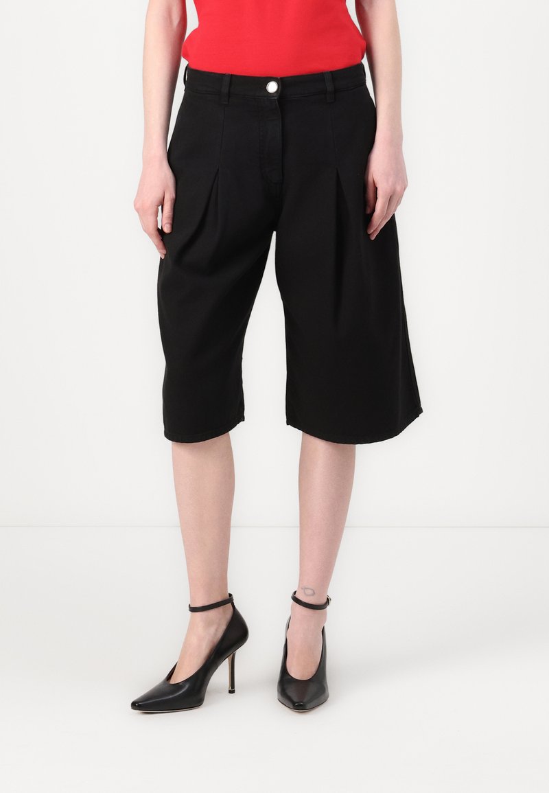 Zwarte capri-broek met geplooide voorkant, zijzakken en knoopsluiting, gecombineerd met zwarte hoge hakken. Glad stofoppervlak.