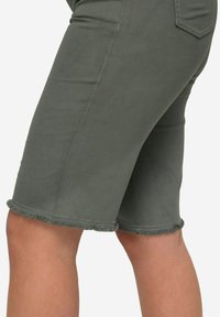 Bermudas de denim verde con dobladillo deshilachado, que presentan un diseño ajustado y bolsillos traseros, destacando una textura suave y un acabado mate.