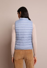 Gilet imbottito azzurro chiaro con cuciture orizzontali, alto colletto e cuciture sulle spalle, abbinato a un maglione beige e pantaloni in corduroy beige.