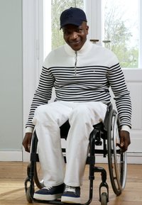 Rolstoelgebruiker zit binnenshuis, draagt een gestreept wit en marineblauw sweater, witte broek en blauwe sneakers. Houten vloeren en lichte muren zijn zichtbaar.