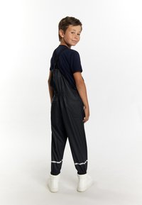 Juoda vandeniui atspari viršutinė apranga su elastingomis rankogaliais, reguliuojamomis petnešomis ir atspindinčiais akcentais, dėvima ant tamsiai mėlynos marškinėlių ir baltų aukštų sportbačių.