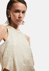 Top senza maniche color beige chiaro con una finitura testurizzata e stropicciata. Il design presenta un collo alto e cade in modo morbido sui lati.
