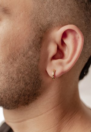 Pendiente de aro de oro con acabado pulido, llevado en la oreja izquierda. La textura es suave, complementando un corte de cabello muy corto y una barba corta.