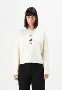 Donna con capelli scuri e lisci che indossa un maglione crema con un ritratto in bianco e nero di una donna con una maglietta nera, abbinato a pantaloni neri.