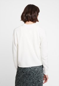 Suéter de punto blanco con canalé y cuello redondo, visto desde atrás, combinado con una falda estampada con estampado de animales en tonos oscuros.