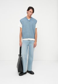 Homem com um colete azul claro sem mangas sobre uma t-shirt branca, jeans azul claro, sapatos pretos, segurando uma grande bolsa tote preta.