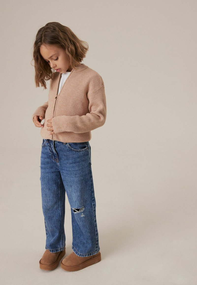 Bež pleten cardigan z zadrgo čez belo majico, v kombinaciji z sproščenimi modrimi kavbojkami z raztrganino na kolenu in rjavimi natikači.