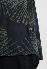 Černá košile s krátkým rukávem, zdobená potiskem palmových listů v zelené a modré barvě, vyrobená z měkké látky, s malou značkovou nášivkou na dolní straně.