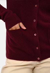 Burgundský cardigan vyrobený z měkké tkaniny, s žebrovanými manžetami, bočními kapsami a světlými knoflíky na přední straně. Kalhoty v neutrální barvě.