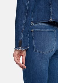 Giubbotto di jeans con polsini sfilacciati e un dettaglio con etichetta nera, abbinato a jeans aderenti coordinati con una tasca posteriore e una leggera scoloritura.