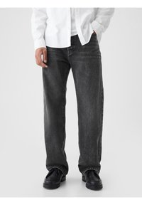 Jean en denim noir à coupe ample, présentant un léger délavage et un design classique à cinq poches. Porté avec des chaussures noires et une chemise blanche.